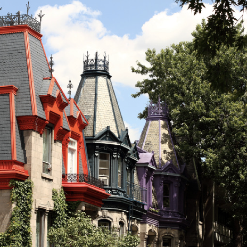 Colorful Victorian-style rooftops of historic townhouses in Le Plateau-Mont-Royal, Montreal, featuring vibrant red, teal, and purple accents under a bright summer sky, partially framed by lush green trees.